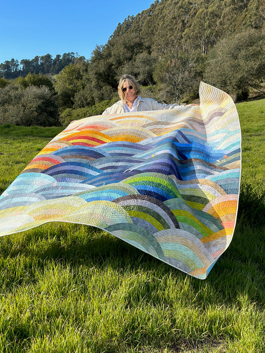 Jennifer Sampou holding the hills quilt outside in a meadow with trees in the background. Hills Quilt kit featuring Sky Ombré Fabric and Chalk and Charcoal fabric, Colorful patterned fabric with circular designs in various shades of blue, green, yellow, and red. Molehills Quilt Pattern by Latifah Saafir.