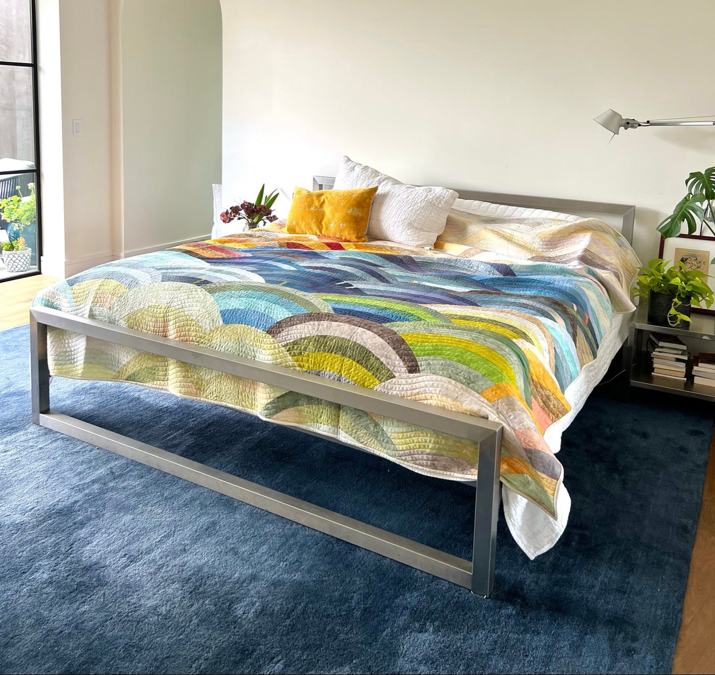 Bed with colorful bedding in a bedroom setting. Hills Quilt kit featuring Sky Ombré Fabric and Chalk and Charcoal fabric, Colorful patterned fabric with circular designs in various shades of blue, green, yellow, and red. Molehills Quilt Pattern by Latifah Saafir.